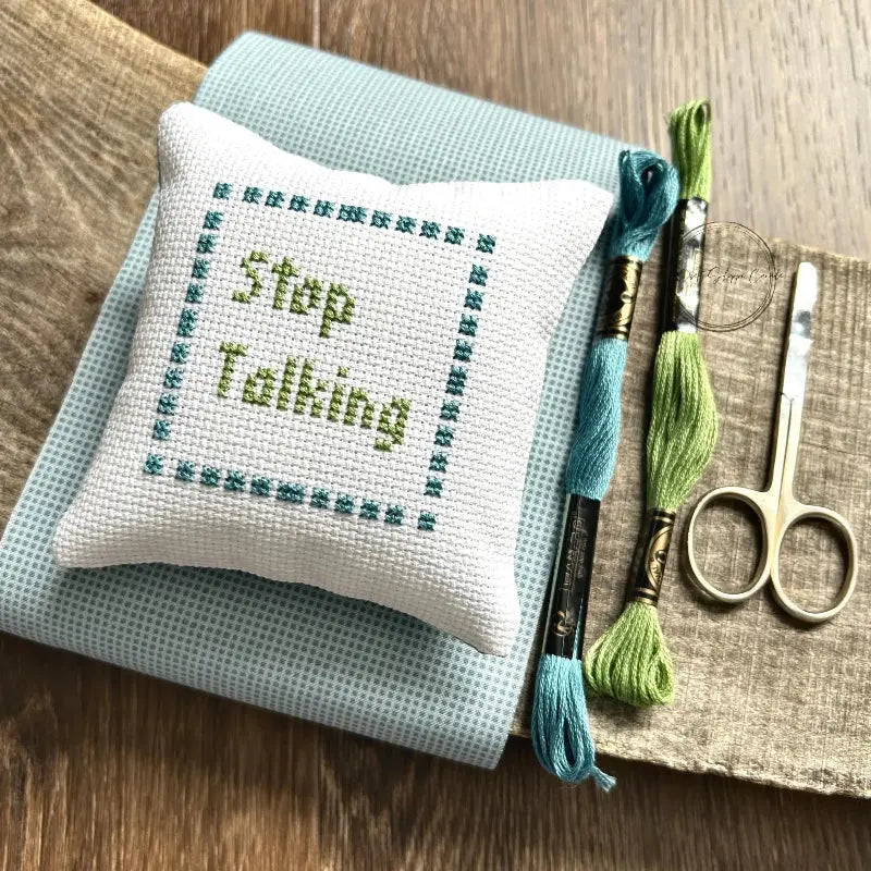 The finished pillow, made from this cross stitch kit, is sitting on a board with all of the supplies needed.