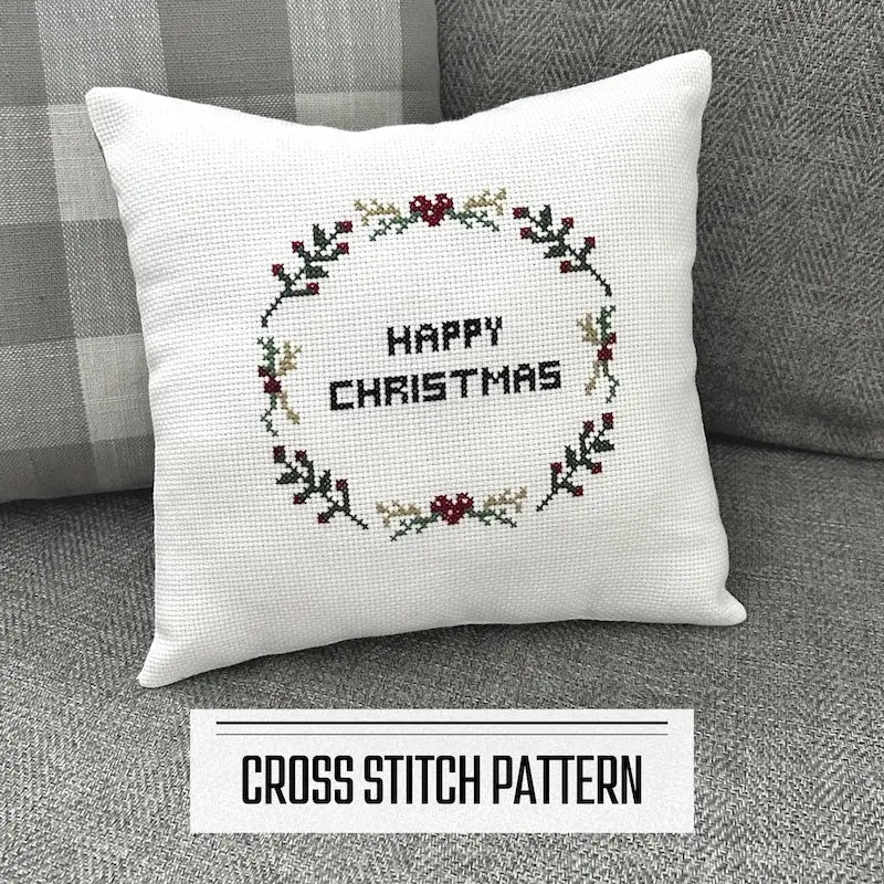 A DIY cross stitch pattern for a Christmas throw pillow. The finished pillow is sitting on a couch against a cushion.