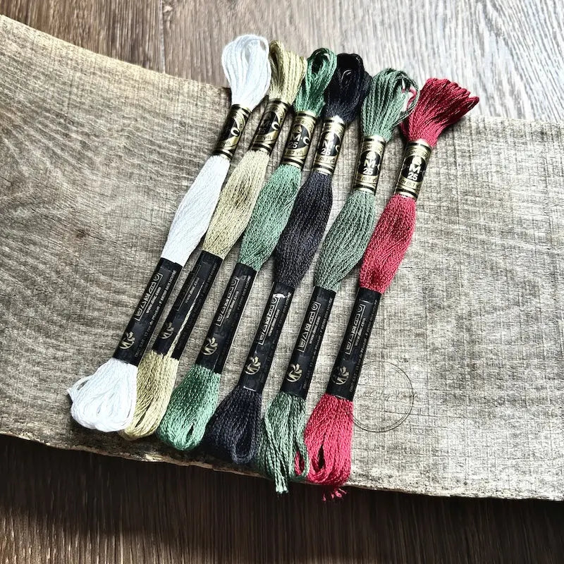 DMC floss, that would be used in this cross stitch pattern, is sitting on a wooden board.