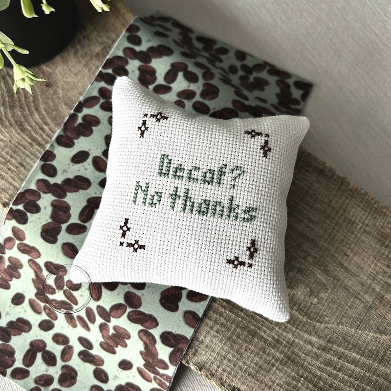 The finished pillow, made from this cross stitch kit, is sitting on a wooden board with a piece of matching fabric.