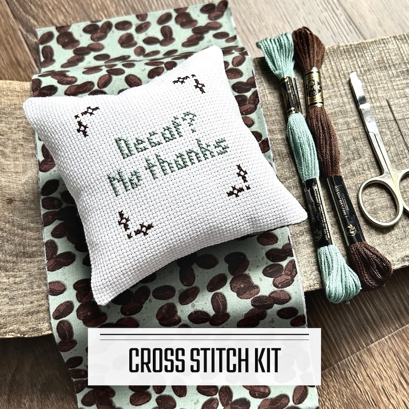 A DIY cross stitch kit for a mini pillow that can be used as a pin cushion. The finished pillow is sitting on a wooden board.
