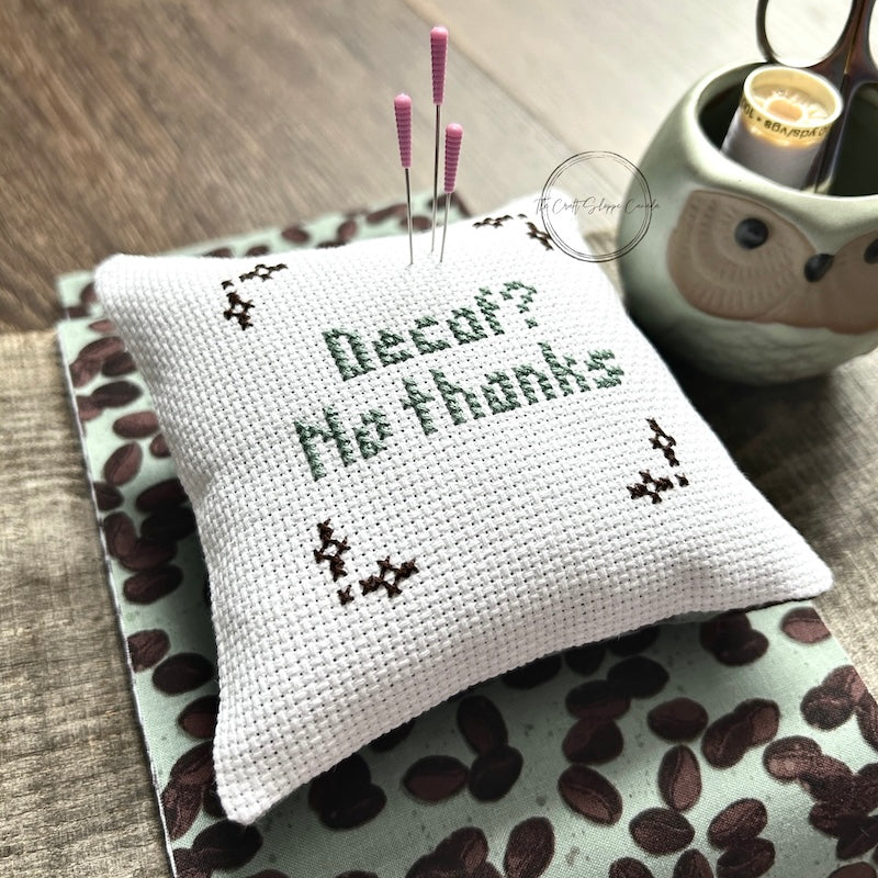 The finished pillow, made from this cross stitch kit, is sitting on a wooden board with pins sticking out.