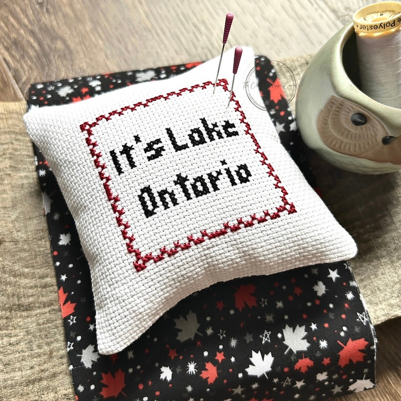 The finished pillow, made from this cross stitch kit, is sitting on a wooden board with pins sticking out.