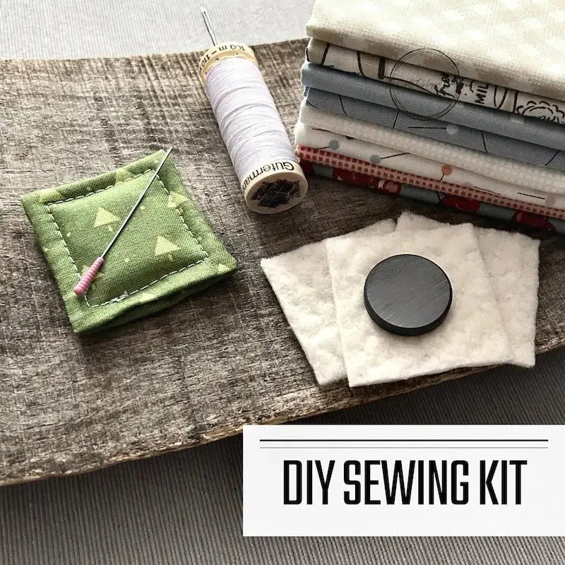 A DIY sewing kit for a needle minder/fridge magnet. There are 2 needle minders lying on a wooden board with fabric, a spool of thread and a magnet.