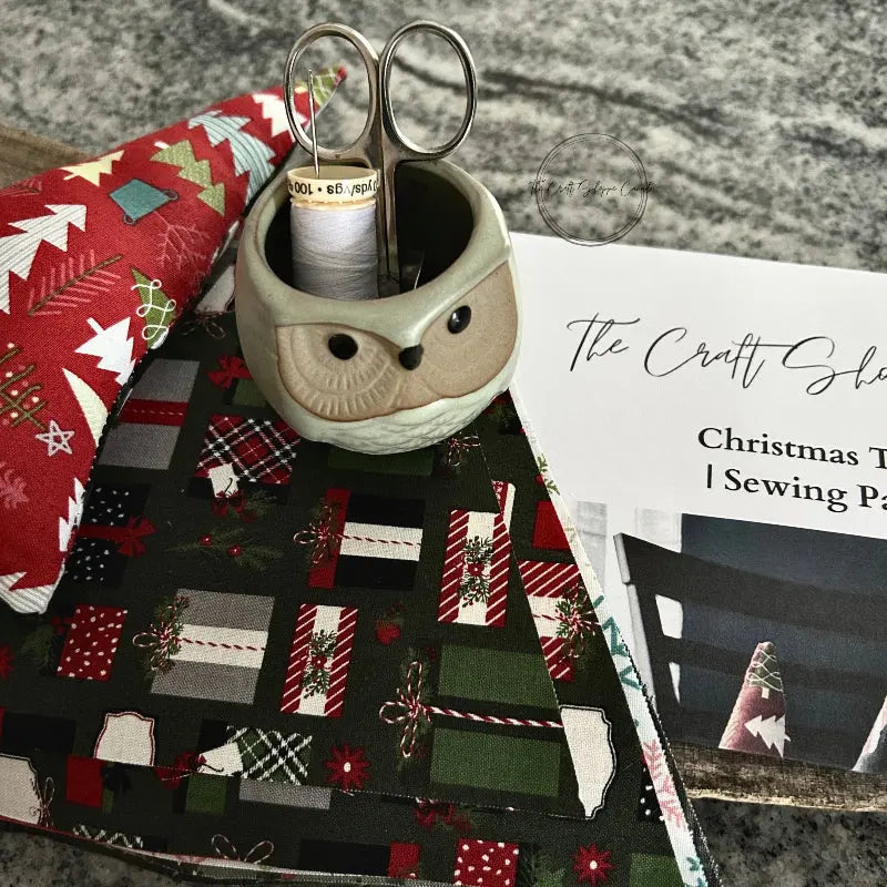 Fabric used to make the Christmas trees, from this sewing pattern, is sitting on a wooden board with a pair of scissors, a spool of thread, and sewing pins.