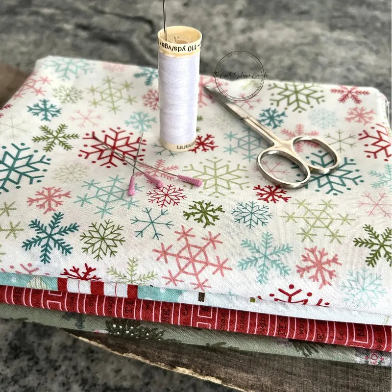 Fabric used to make these Christmas trees, from this sewing pattern, is sitting on a wooden board with a pair of scissors, a spool of thread, and sewing pins.