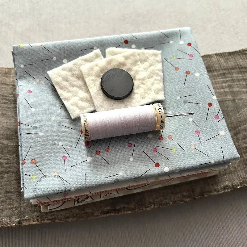 A stack of fabric, cotton batting and a magnet, that would be used in this sewing pattern, are sitting on a wooden board with a spool of thread and a hand sewing needle.