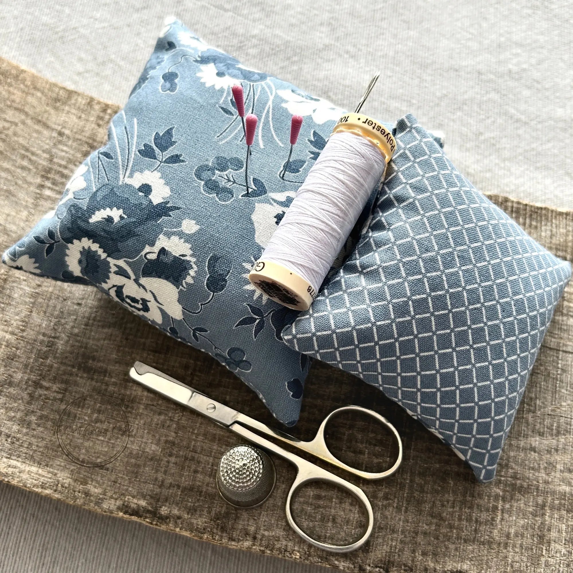 The finished pin cushions, made from this sewing pattern, are sitting on a wooden board with scissors, a spool of white thread and a hand sewing needle.