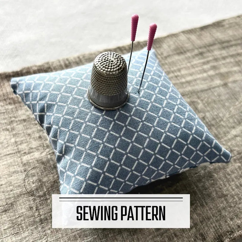 A digital PDF sewing pattern for a pin cushion. The pin cushion is sitting on a wooden board with sewing pins stuck on top with a thimble.