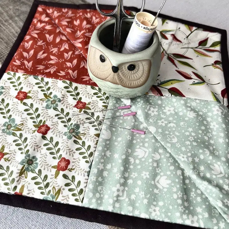 The pot holder trivet, made from this sewing pattern, is sitting on a wooden board with a pair of scissors, a spool of thread, and sewing pins.