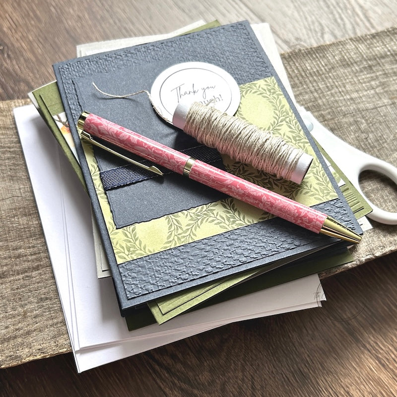 A finished stack of greeting cards, made from a card making kit, sitting on a wooden board with a pen and a roll of twine lying on top.