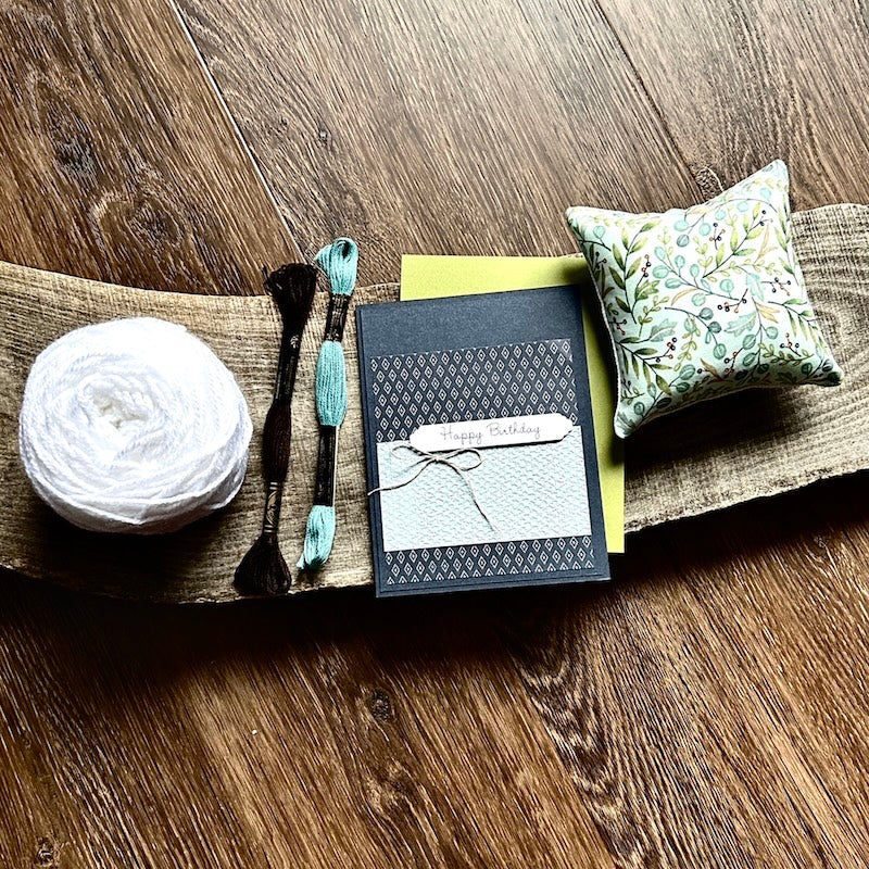 A ball of yarn, greeting cards, a pin cushion and DMC floss is sitting on a wooden board to promote the gift card.