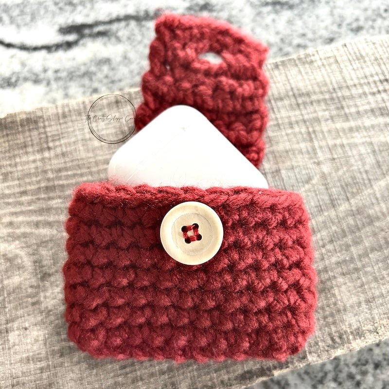 The red ear bud case with a pair of ear buds poking out of the top, laying on a wooden board - The Craft Shoppe Canada