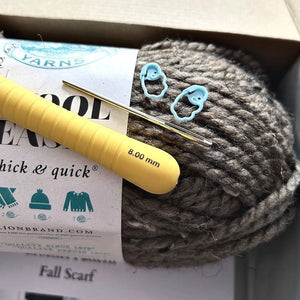 A ball of brown yarn in a box with a crochet hook, a tapestry needle and 2 stitch markets on it - The Craft Shoppe Canada