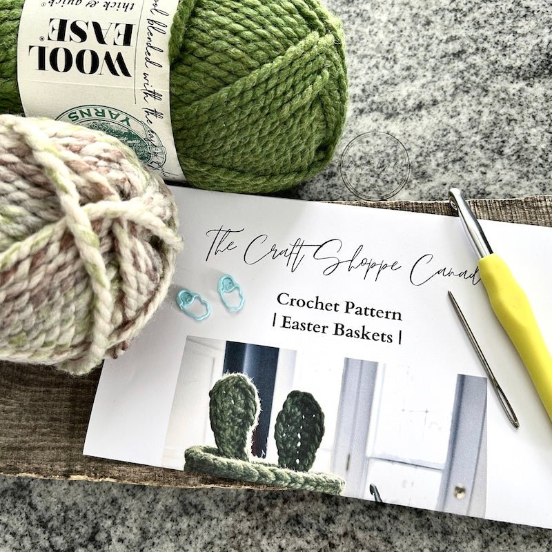 A copy of the pattern laying on a wooden board with yarn, a crochet hook, a tapestry needle and 2 stitch markers laying on top - The Craft Shoppe Canada