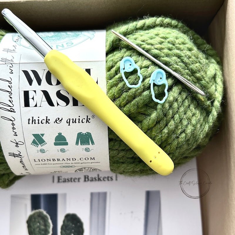 A crochet pattern for an Easter bunny basket set. The baskets are sitting on a wooden board with yarn and a crochet hook sitting in the smaller basket - The Craft Shoppe Canada