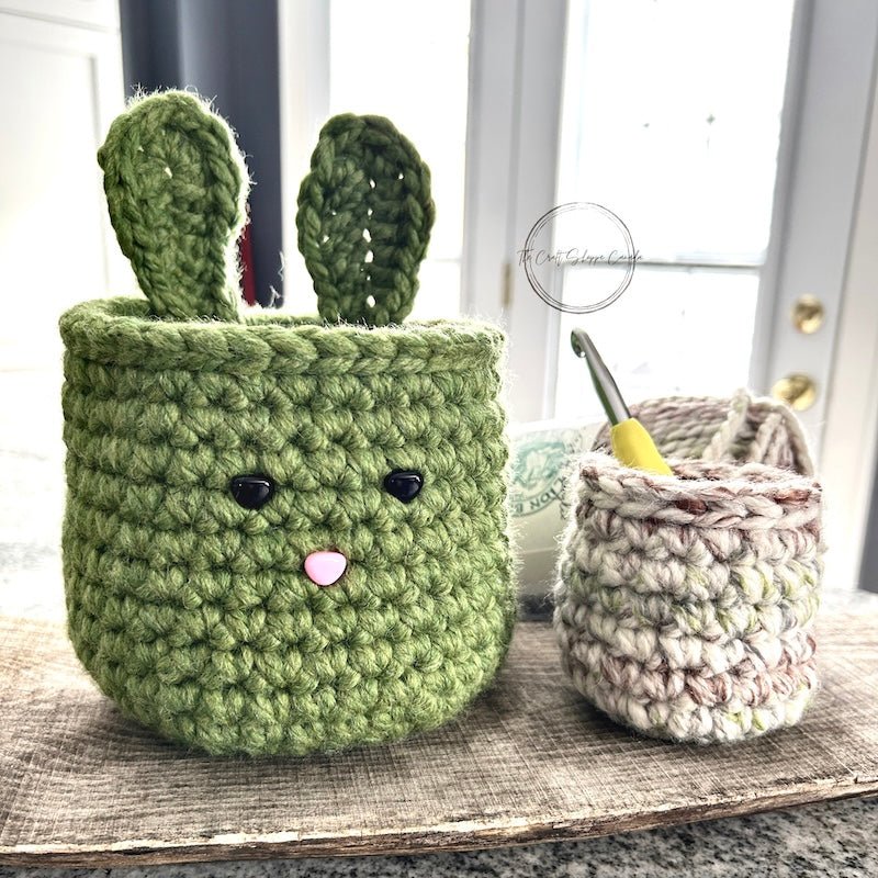 The crocheted Easter basket set sitting on a wooden board with yarn and a crochet hook sitting in the smaller basket - The Craft Shoppe Canada