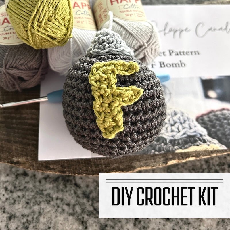 A DIY crochet kit for an amigurumi plush F bomb. The F bomb is sitting on a wooden board with a crochet hook on top of the pattern - The Craft Shoppe Canada