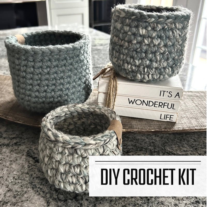 A crochet kit for a set of 3 baskets. The baskets are sitting on a wooden board on a counter - The Craft Shoppe Canada