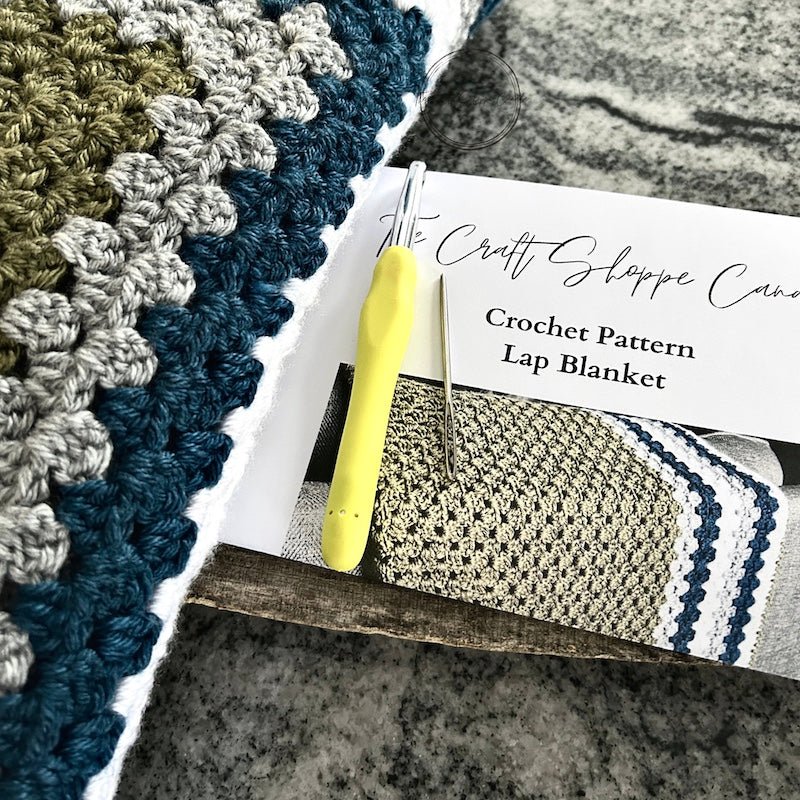 The finished blanket laying on a wooden board, over the crochet pattern with a crochet hook and a tapestry needle - The Craft Shoppe Canada