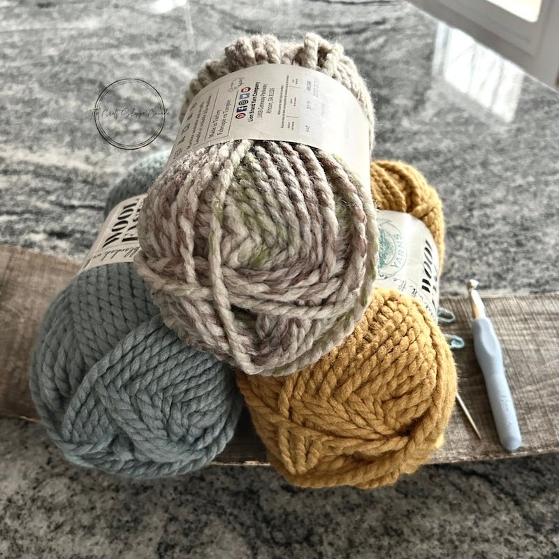 There are 3 balls of yarn sitting on a wooden board with a crochet hook. The yarn colours are blue, yellow and a green/ivory mix - The Craft Shoppe Canada