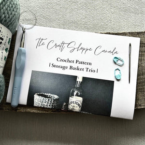 A copy of the pattern laying on a wooden board with yarn, a crochet hook, stitch markers and a tapestry needle laying on top - The Craft Shoppe Canada