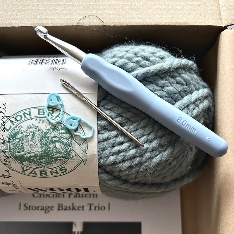 A crochet kit for a set of 3 baskets. The baskets are sitting on a wooden board on a counter - The Craft Shoppe Canada