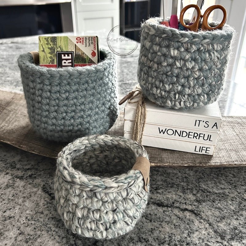 The 3 baskets from this crochet kit are sitting on a wooden board with tea and and craft supplies inside to show how they can be used - The Craft Shoppe Canada