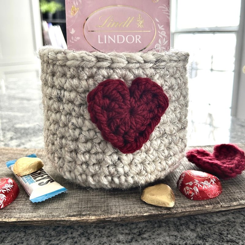 The crocheted heart basket sitting on a wooden board with chocolates inside and out of the basket - The Craft Shoppe Canada