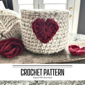 A crochet pattern for a Valentine heart basket. The basket is sitting on a wooden board with yarn and a red heart beside it - The Craft Shoppe Canada