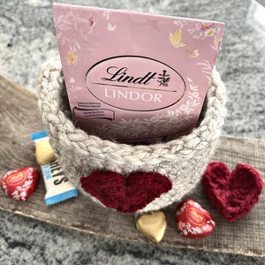 The crocheted heart basket sitting on a wooden board with chocolates inside and out of the basket - The Craft Shoppe Canada