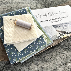 The sewing pattern is sitting on a wooden board beside a pile of fabric, a spool of white thread and a sewing needle - The Craft Shoppe Canada