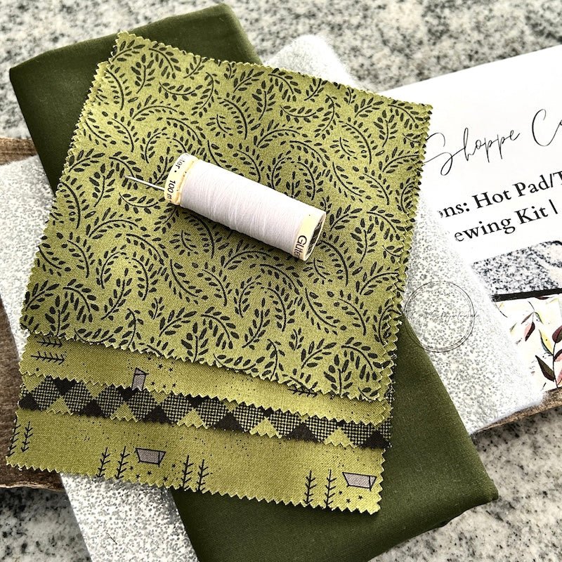 A DIY sewing kit for a pot holder. The pot holder is sitting on a wooden board with a pair of scissors, a thimble, and sewing pins - The Craft Shoppe Canada