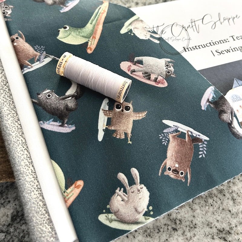 The sewing pattern is sitting on a wooden board beside a pile of fabric, a spool of white thread and a sewing needle - The Craft Shoppe Canada