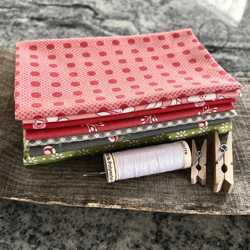 A stack of fabric, a spool of thread and a sewing needle laying on a wooden board - The Craft Shoppe Canada