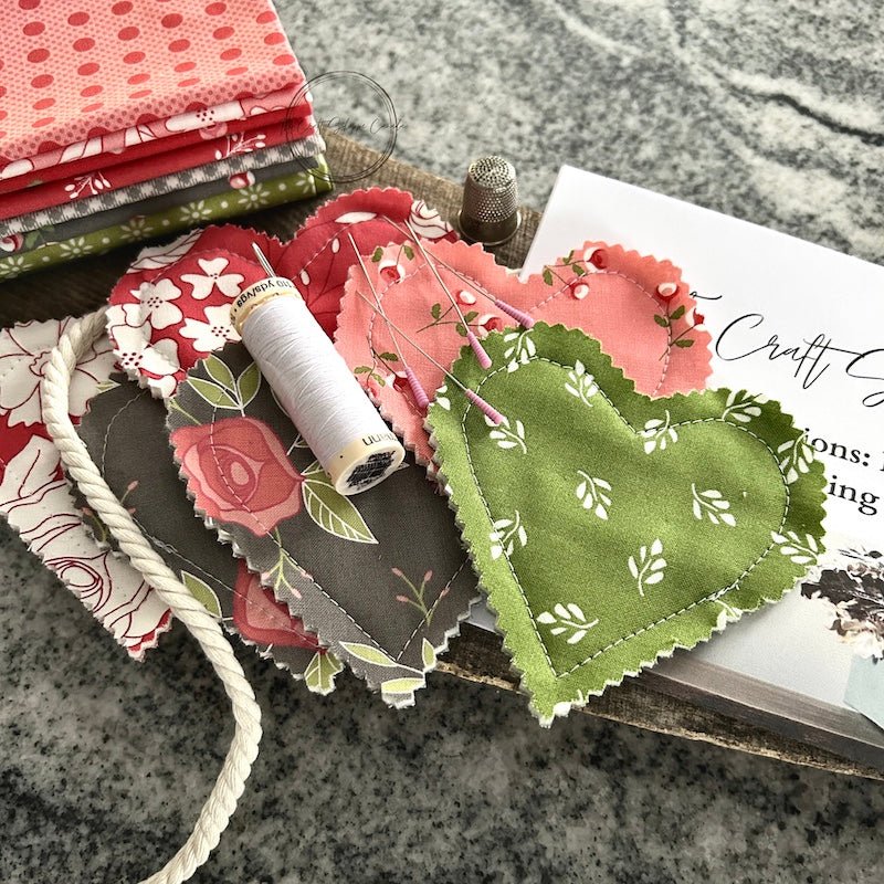 The sewing pattern with finished hearts, sewing pins, fabric, a spool of thread and a sewing needle - The Craft Shoppe Canada
